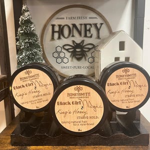May include: Three black jars of hair product, labeled "Black Girl Magic" and "King's Honey," sit on a wooden shelf. The jars are 6 oz each. A sign in the background reads "Farm Fresh Honey" with a bee graphic.