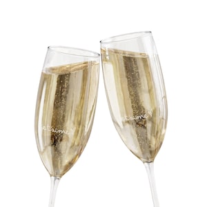 Puede incluir: Dos copas de champán chocando, llenas de líquido dorado brillante. Las copas tienen la inscripción "je t'aime". El fondo es blanco.