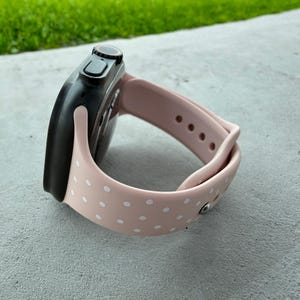 Op de afbeelding: Een zwarte smartwatch met een blush roze siliconen band met witte stippen. Het horloge staat schuin, waardoor het scherm en het bandontwerp te zien zijn. De band is vastgezet met een zilveren sluiting. De achtergrond is een grijs oppervlak met groen gras.