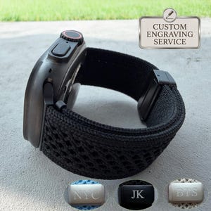 May include: A black smartwatch with a textured, woven nylon band featuring a black metal clasp. A rectangular sign above displays the text CUSTOM ENGRAVING SERVICE. Three small metal plates below show the engraved letters NYC, JK, and BTS.