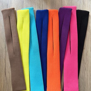 May include: A selection of vibrantly coloured ribbed knit leg warmers. The colours include brown, yellow, turquoise, blue, orange, purple, pink, and black. Each leg warmer features a vertical slit. The items are arranged side by side.