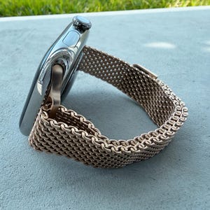 Puede incluir: Un reloj inteligente plateado con una correa de malla de oro rosa. La esfera del reloj es oscura con forma rectangular redondeada. La correa está hecha de un material metálico tejido y se abrocha con un cierre.