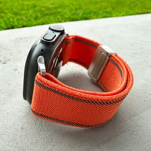 Puede incluir: Un reloj inteligente negro con una correa a rayas naranja y gris. La correa está hecha de un material tejido y tiene un cierre plateado. El reloj está sobre una superficie gris con hierba verde en el fondo.