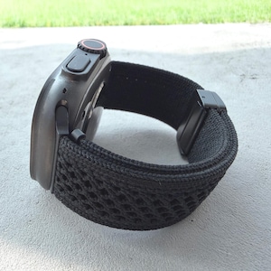 May include: A black smartwatch with a black woven band. The watch face is dark with a red accent on the dial. The band has a unique open weave design and a black clasp.