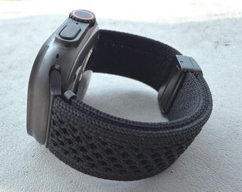 May include: A black smartwatch with a black woven band. The watch face is dark with a red accent on the dial. The band has a unique open weave design and a black clasp.