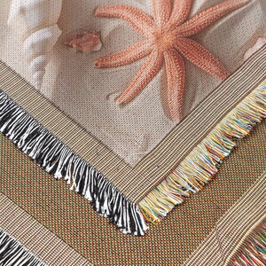 May include: A woven textile featuring a starfish, seashells, and sand-like texture. The border has a brown woven pattern with black, and multicolored fringe. The design evokes a beach theme.
