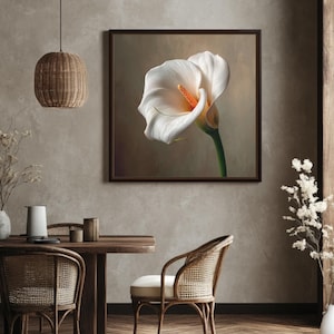 Op de afbeelding: Een ingelijste print van een witte calla met een oranje spadix. De bloem staat tegen een zachte, neutrale achtergrond. De print wordt getoond in een eetkamer met een houten tafel en stoelen.
