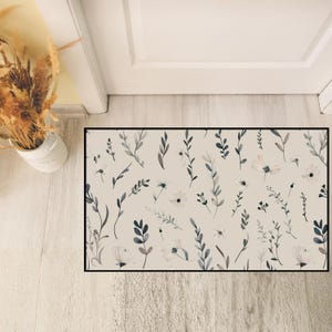 May include: A rectangular beige doormat with a black border. The mat features a watercolor floral pattern of white flowers and gray-blue leaves and stems. The design is repeated across the mat's surface.