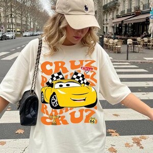 A woman wearing a white t-shirt with a cartoon car graphic and a cap is standing on a city street.