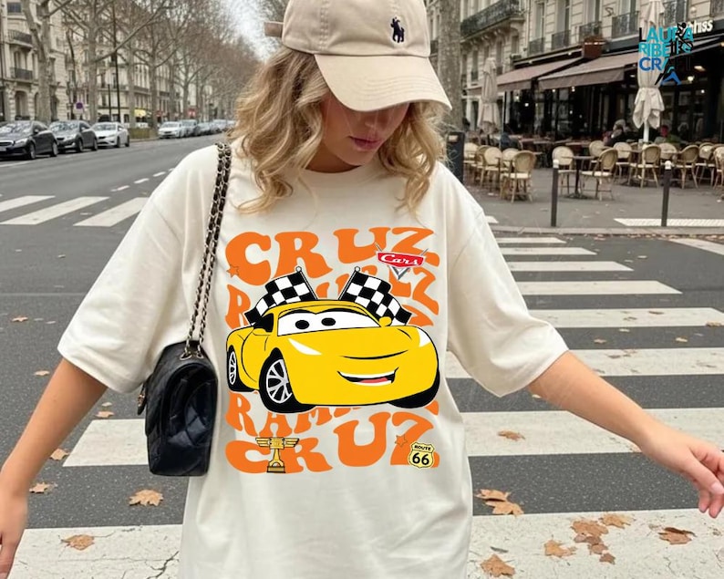 A woman wearing a white t-shirt with a cartoon car graphic and a cap is standing on a city street.