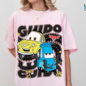 A pink t-shirt with a graphic design featuring two cartoon cars, one yellow and one blue, along with the text "GUIDO" and "Cars" printed on it.