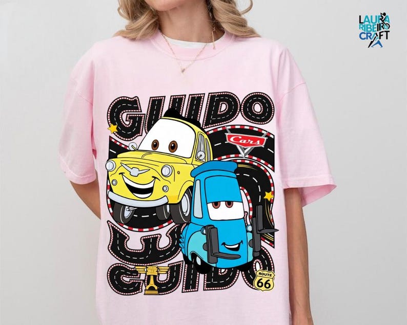 A pink t-shirt with a graphic design featuring two cartoon cars, one yellow and one blue, along with the text "GUIDO" and "Cars" printed on it.