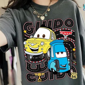 A person wearing a gray t-shirt with a graphic design featuring two cartoon cars, one yellow and one blue, along with the text "GUIDO GUIDO" and "Cars".