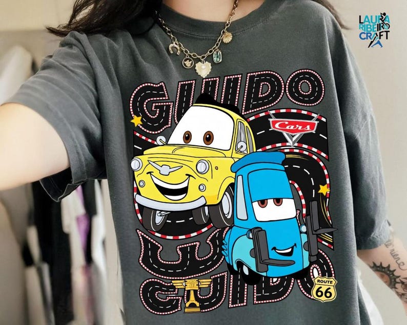 A person wearing a gray t-shirt with a graphic design featuring two cartoon cars, one yellow and one blue, along with the text "GUIDO GUIDO" and "Cars".