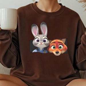 A person is sitting on a couch, wearing a brown sweater with a graphic of two cartoon rabbit characters, one gray and one orange, and holding a white mug.