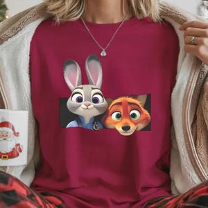 A person wearing a red shirt with a graphic of two cartoon animals, a rabbit and a fox, peeking out from behind a mug with a Santa Claus design.