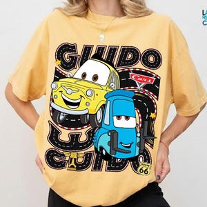 A yellow t-shirt with a graphic design featuring two cartoon cars, one yellow and one blue, along with the text "GUIDO GUIDO" and "Cars" written on it.