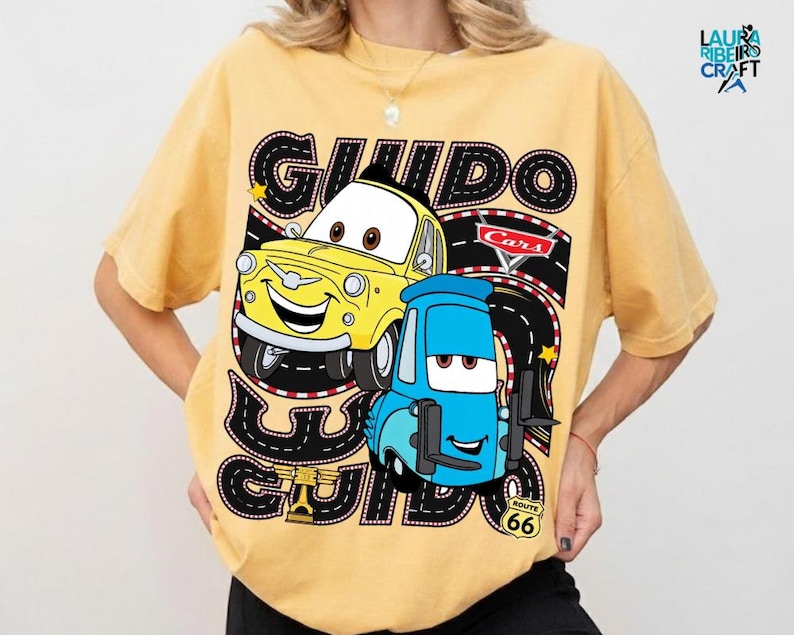 A yellow t-shirt with a graphic design featuring two cartoon cars, one yellow and one blue, along with the text "GUIDO GUIDO" and "Cars" written on it.