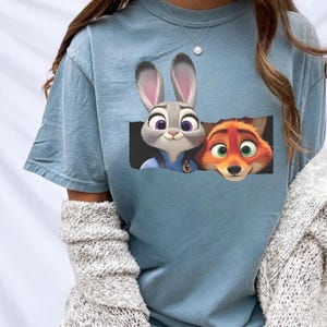 A person wearing a blue t-shirt with a graphic of a rabbit and a fox peeking out from a window.