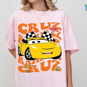 A person wearing a pink t-shirt with a graphic of a yellow race car and racing-related text.