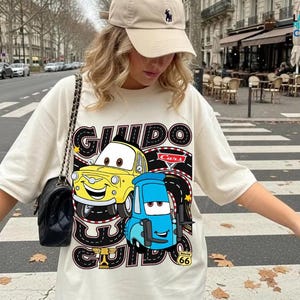 A woman wearing a white t-shirt with a graphic of two cartoon cars, one yellow and one blue, standing on a street.