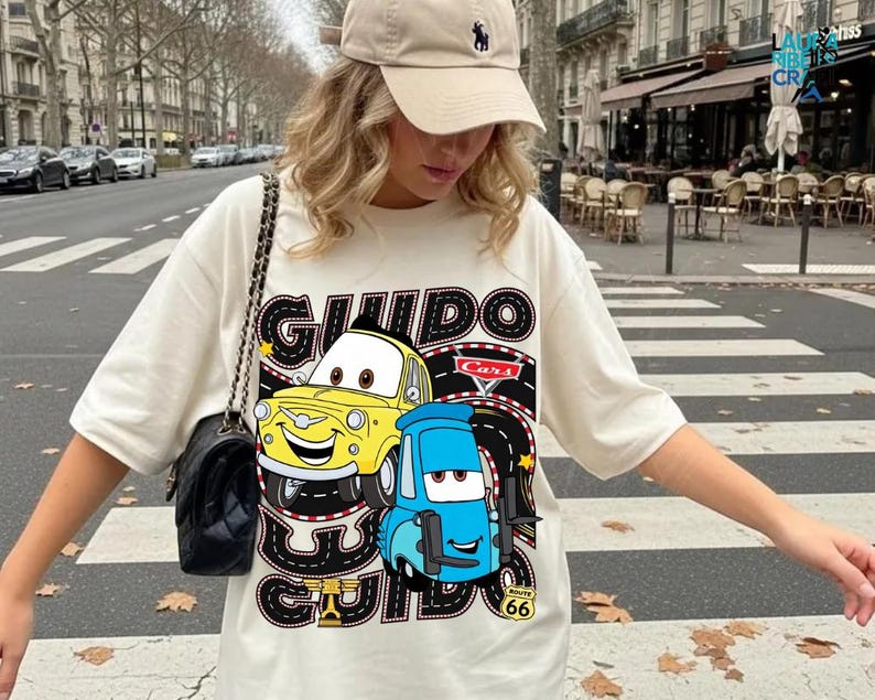 A woman wearing a white t-shirt with a graphic of two cartoon cars, one yellow and one blue, standing on a street.