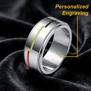 May include: A silver-toned metal spinner ring featuring three thin, vertical lines in red, yellow, and green. The ring has a smooth, polished finish and includes the text Personalized Engraving with an arrow pointing to the inner band.