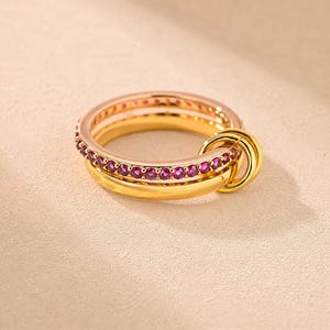 Op de afbeelding: Een gouden ring met een band bezet met een rij kleine, ronde, felroze edelstenen. De ring is verbonden met twee kleinere, gladde, in elkaar grijpende gouden ringen die aan één kant van de hoofdring hangen voor een gelaagd ontwerp.