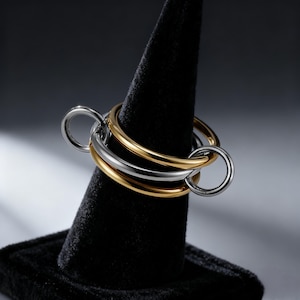 Op de afbeelding: Een multi-band ring met een modern design. De ring heeft drie banden: één in goud, één in zilver en één in goud. De banden zijn met elkaar verbonden met twee zilveren ringen. De ring wordt gepresenteerd op een zwarte fluwelen ringhouder.