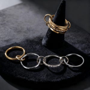 May include: An assortment of gold and silver rings. Some rings are adorned with small, sparkling stones. One set of rings is displayed on a black cone-shaped stand. The rings are linked together in various configurations.