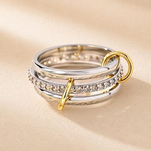 以下が含まれることがあります： 2本の滑らかなシルバー調のバンドと、透明な石が埋め込まれた中央のバンドで構成されたマルチバンドリング。2つの小さな磨かれたゴールド調のリングがバンドに通されており、それらを重ね合わせた円形のデザインで繋いでいます。