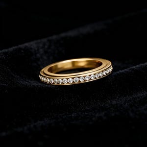 Puede incluir: Un anillo dorado con una fila de pequeñas piedras preciosas transparentes. El anillo se muestra sobre un fondo oscuro, resaltando su acabado pulido y el brillo de las piedras. El diseño es simple y elegante, adecuado para diversas ocasiones. El diámetro del anillo es de unos 2 cm.