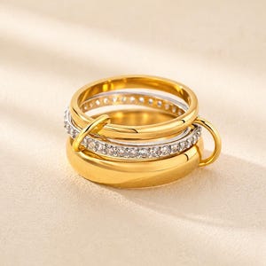 May include: A multi-layered ring set featuring a thick gold-tone band, a central silver-tone band encrusted with clear stones, and a smooth gold-tone band, all interconnected by a small gold-tone loop attached to the side of the rings.
