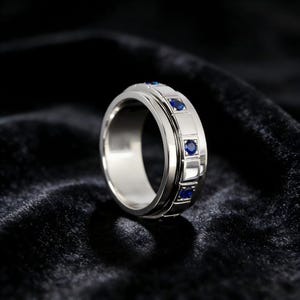 May include: A silver ring with a series of rectangular and square accents, each containing a small, round, blue gemstone. The ring is set against a dark, textured background, highlighting its polished metal and vibrant blue stones.