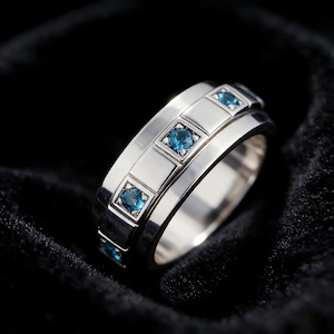 Könnte beinhalten: Ein silberner Ring mit einem Band mit quadratisch gefassten, blauen Edelsteinen. Der Ring hat eine polierte Oberfläche und ist vor einem dunklen, samtigen Hintergrund platziert, der das Design und die Farbe der Edelsteine hervorhebt.