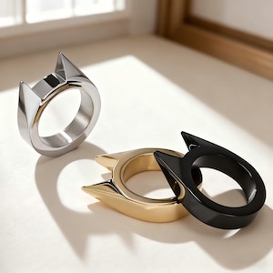 Könnte beinhalten: Drei Katzenohr-Ringe in Silber, Gold und Schwarz. Die Ringe haben ein minimalistisches Design mit spitzen Ohren. Die Ringe werden auf einer weißen Oberfläche präsentiert, die von natürlichem Licht beleuchtet wird.