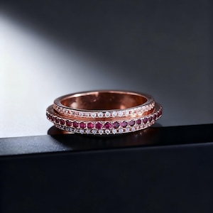 May include: Rose gold ring with two bands. The top band is lined with small white stones, while the bottom band is lined with small red stones. The ring is set against a dark background.