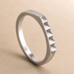 May include: A silver-toned metal ring featuring a smooth, rounded band that transitions into a flat, rectangular section on one side. This section is adorned with four distinct, evenly spaced, pointed triangular spikes protruding outward.