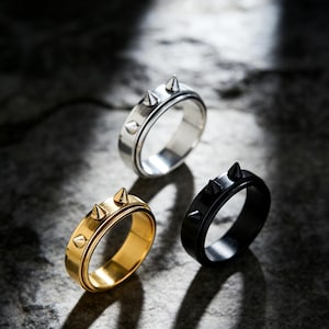 May include: Three rings with a spiked design. One ring is silver, one is gold, and the third is black. Each ring has three evenly spaced spikes. The rings are displayed on a dark, textured surface.
