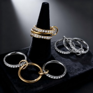 Può includere: Un assortimento di anelli in oro e argento, alcuni adornati con piccole pietre scintillanti. Diversi anelli sono collegati tra loro, alcuni esposti su un cono di velluto nero. Gli anelli risaltano su uno sfondo scuro.