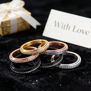 Op de afbeelding: Een assortiment ringen in goud, roségoud, zwart en zilver, elk met een band van sprankelende stenen. Een geschenkdoos met een wit lint en een kaart met de tekst "With Love" staan op de achtergrond.