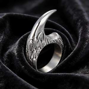 May include: A silver-toned metal ring featuring a prominent, curved claw or talon design. The band is textured with detailed, overlapping feather-like engravings that wrap around the finger opening, creating a sharp, pointed silhouette.