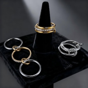 May include: An assortment of rings in gold and silver tones, some adorned with small, sparkling stones. One gold ring is displayed on a black cone, while others are arranged on a black velvet surface, showcasing various designs and styles.