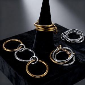 Peut inclure: Plusieurs bagues dorées et argentées, certaines entrelacées, présentées sur une surface noire. Un ensemble de bagues est enroulé autour d'un cône noir. Les designs des bagues varient, mettant en valeur un style de bijoux moderne.