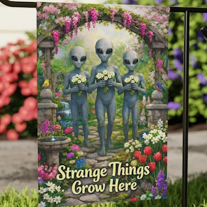 Puede incluir: Una bandera de jardín con tres extraterrestres grises sosteniendo ramos de flores blancas, en un jardín con un arco. La bandera tiene el texto "Strange Things Grow Here" y está rodeada de flores coloridas.