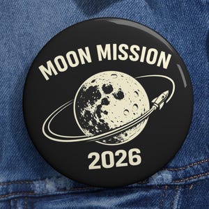 May include: A black button with the words "MOON MISSION" in white, above a detailed illustration of the moon with a rocket ship circling it. The year "2026" is printed below the moon illustration.