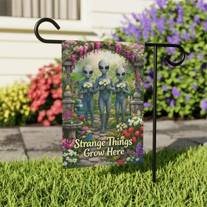 May include: A rectangular garden flag hanging on a black metal stand features three gray aliens standing on a stone path, each holding a bouquet of white flowers. The text Strange Things Grow Here is printed in yellow at the bottom of the flag.