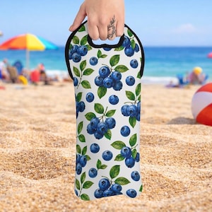 Peut inclure: Un sac fourre-tout blanc pour bouteille de vin avec un motif de myrtilles et de feuilles vertes. Le sac a une poignée et une bordure noires. L'arrière-plan est une plage de sable avec un parasol coloré et l'océan.