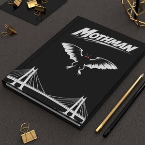 May include: Black journal with the word "Mothman" in white, along with a white graphic of a winged creature with red eyes. A white bridge graphic is on the bottom left. Gold and black pens and gold binder clips are on the dark gray surface.