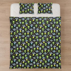 Peut inclure: Une parure de lit sur fond noir avec un motif répétitif de têtes d'extraterrestres vertes, de soucoupes volantes grises et de petites étoiles jaunes. L'ensemble comprend une grande couette rectangulaire et deux oreillers carrés assortis.